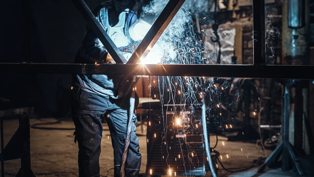 Soldador utilizando máscara de proteção realiza soldagem em estrutura metálica com faíscas e luz intensa no ambiente industrial.